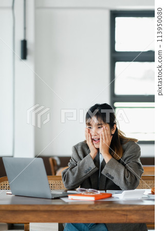 Woman in a professional setting looks astonished, hands framing her face, as she reacts to what she sees on her laptop screen. 113950080