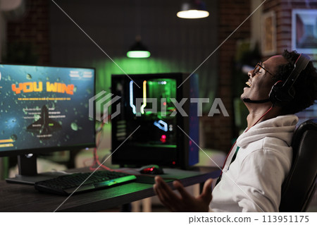 Excited gamer celebrating winning online multiplayer futuristic videogame match. African american man enjoying leisure time at home, feeling ecstatic about gaming championship victory 113951175