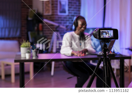 Focus shot on modern camera recording vlogger in blurry background talking about family and relationships subjects. Woman producing online show using high quality equipment in rgb lights studio 113951182
