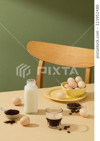 A beige table with a glass bottle of milk, a glass of coffee, some eggs on a yellow bowl lay on. Decorated by eggs, some coffee beans and powdered coffee. Copy space and blank space for designing 113952480