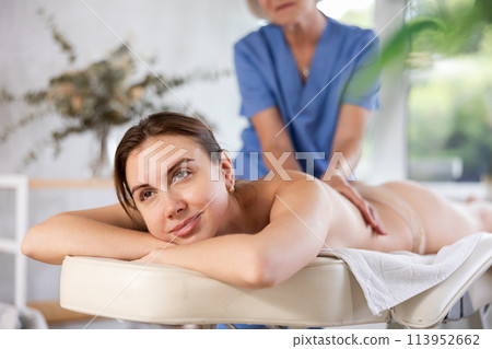 Cropped mature woman actively rubs lower back of young woman during general massage procedure. Cropped mature woman actively rubs lower back of young woman during general massage procedure. 113952662