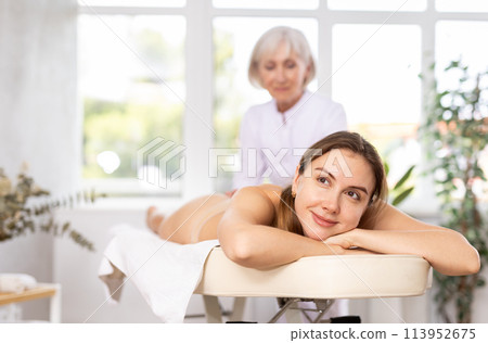 Blurred mature woman actively rubs lower back of young woman during general massage procedure. 113952675