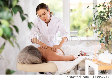 Unrecognizable elderly woman lies face down on couch and enjoy spa treatments, shoulder massage Unrecognizable elderly woman lies face down on couch and enjoy spa treatments, shoulder massage 113952717