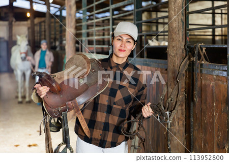 Female rancher carrying horse tack and saddle in horse farm Female rancher carrying horse tack and saddle in horse farm 113952800