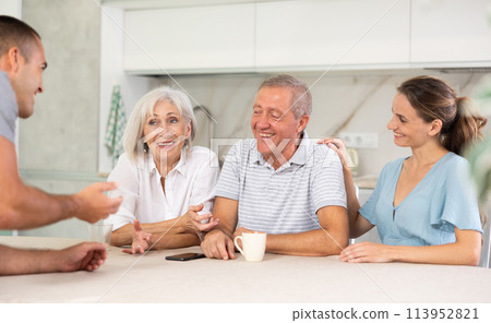 Smiling parents and adult children sit at the table with tea and talk about home life 113952821