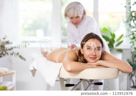Vague mature woman actively rubs buttocks of young woman during general massage procedure. Vague mature woman actively rubs buttocks of young woman during general massage procedure. 113952876