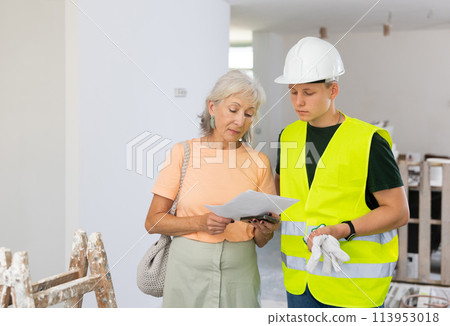 Mature woman discussing room renovation work plan with guy in protective helmet anf yellow vest 113953018