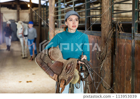 Woman horse breeder carrying leather saddle Woman horse breeder carrying leather saddle 113953039
