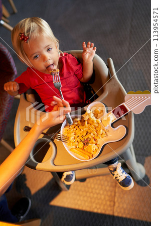 Mom feeds a little girl pasta with meat from a plate in the shape of a guitar. High quality photo Mom feeds a little girl pasta with meat from a plate in the shape of a guitar. High quality photo 113954571