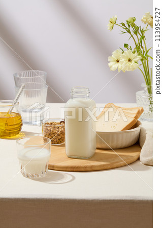 Unlabeled bottle of milk, a cup of soybeans and a bowl containing sandwiches placed on a cutting board. A jar filled with honey and a flower pot decorated. Branding mockup 113954727