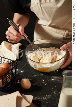 A baker wearing apron is using a whisk to mix eggs and flour inside a glass bowl. Black table featured a basket of eggs, sandwiches and a bottle of milk 113954756