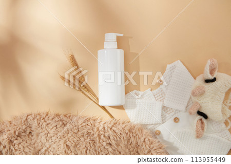 Milk brown background against the layout for baby products with a bottle without label, decorated by clothes for baby, brown feather carpet and a wheat branch. Mock up for advertising, front view 113955449
