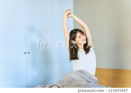 A woman in her 30s stretches her body in a good mood after waking up in her bedroom at home A woman in her 30s stretches her body in a good mood after waking up in her bedroom at home 113955937