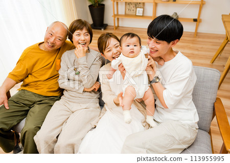 A large family of five people, consisting of three generations: a newborn baby, parents, and senior grandfather and grandmother, all sitting together on a sofa in the living room A large family of five people, consisting of three generations: a newborn baby, parents, and senior grandfather and grandmother, all sitting together on a sofa in the living room 113955995