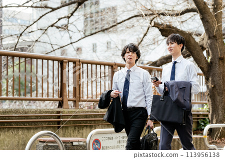 Two young businessmen walking side by side and talking with smartphones along a green road lined with trees in Nakameguro 113956138