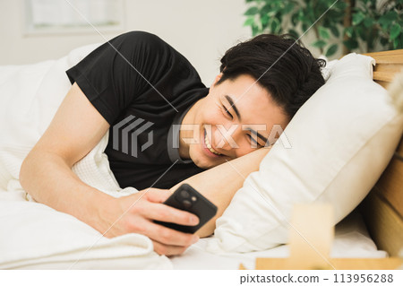A Japanese man in his 20s or 30s operating a smartphone while lying on his bed in a room 113956288