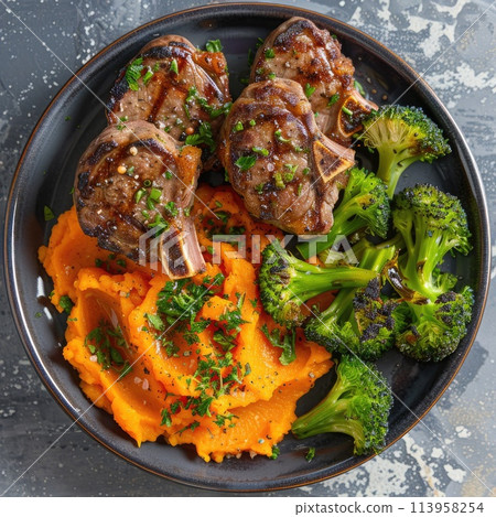 Roasted lamb chops with a side of mashed sweet potato and steamed vegetables. The steamed vegetables are a mix of broccoli and carrots. 113958254