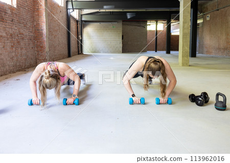 Two Women Doing Push-Ups with Dumbbells in a Spacious Industrial Gym 113962016