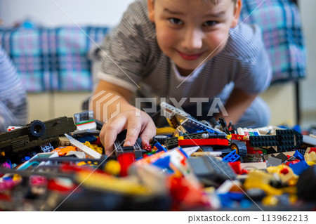 A cute child assembles a car from a construction set. Sitting on the floor, the child searches in a large pile of construction kit for the necessary part for his toy. Constructor concept. A cute child assembles a car from a construction set. Sitting on the floor, the child searches in a large pile of construction kit for the necessary part for his toy. Constructor concept. 113962213