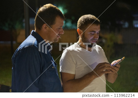 Two friends look at the phone in the park on a warm summer night. Friends walking through the park at night in summer. A man shows something on his phone to his friend while standing in the park. Two friends look at the phone in the park on a warm summer night. Friends walking through the park at night in summer. A man shows something on his phone to his friend while standing in the park. 113962214