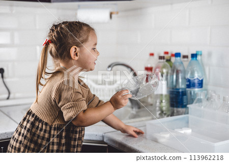 Beautiful girl child puts a plastic bottle in a recycling box. A child takes part in sorting plastic for recycling. The family teaches children to care about nature and recycle plastic. 113962218