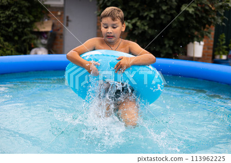 A child boy in an inflatable ring splashes in the pool. On a sunny summer day, a child boy swims in an inflatable pool in the courtyard of his house. The child has fun in the pool. 113962225