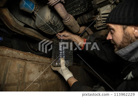 The mechanic removes the plug from the tank, draining the old engine oil from the engine. A mechanic holds a container for draining engine oil to replace it with new one. Machine maintenance. The mechanic removes the plug from the tank, draining the old engine oil from the engine. A mechanic holds a container for draining engine oil to replace it with new one. Machine maintenance. 113962229