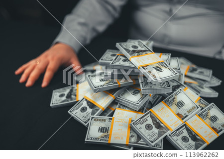 Side view of a large pile of stacks of dollars on a black background next to a seated man. The man made a lot of money. Wealth in front of a man in the form of wads of money. Side view of a large pile of stacks of dollars on a black background next to a seated man. The man made a lot of money. Wealth in front of a man in the form of wads of money. 113962262