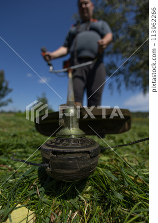 Vertical. On a clear day, a man mows the grass in his backyard with a trimmer. Hold the trimmer firmly. a man mows tall, thick grass in his backyard. A man tidies up the lawn behind his house. 113962266
