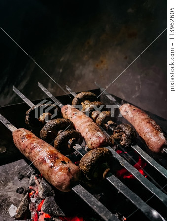 Sausages and mushrooms on skewers over hot coals of a grill Sausages and mushrooms on skewers over hot coals of a grill 113962605