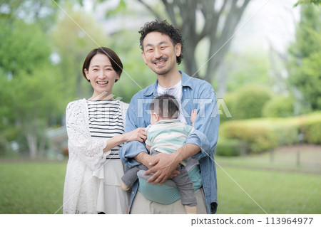 A middle-class family smiling while holding a baby outdoors in fresh greenery 113964977