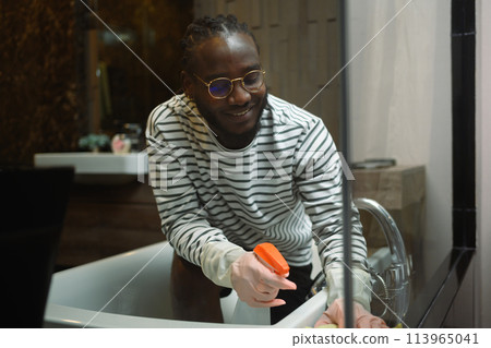 Happy man in rubber gloves cleaning bathtub and faucet in a modern bathroom. Housekeeping concept Happy man in rubber gloves cleaning bathtub and faucet in a modern bathroom. Housekeeping concept 113965041