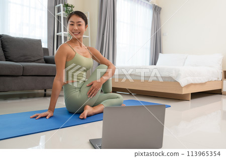 Young happy woman practicing yoga, sitting in Ardha Matsyendrasana exercise or Half lord of the fishes pose on mat in bedroom. Asian woman looking laptop practice yoga online course at home. Young happy woman practicing yoga, sitting in Ardha Matsyendrasana exercise or Half lord of the fishes pose on mat in bedroom. Asian woman looking laptop practice yoga online course at home. 113965354