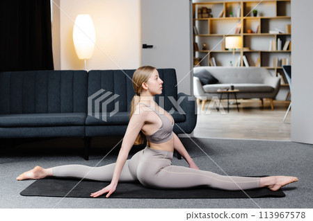 A cute woman in sportswear is sitting in twine on the floor in living room. Healthy lifestyle, yoga, fitness A cute woman in sportswear is sitting in twine on the floor in living room. Healthy lifestyle, yoga, fitness 113967578