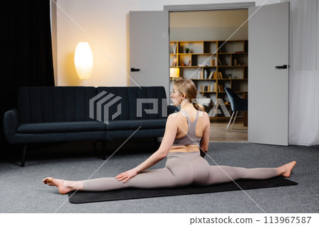 A cute woman in sportswear is sitting in twine on the floor in living room. Healthy lifestyle, yoga, fitness 113967587