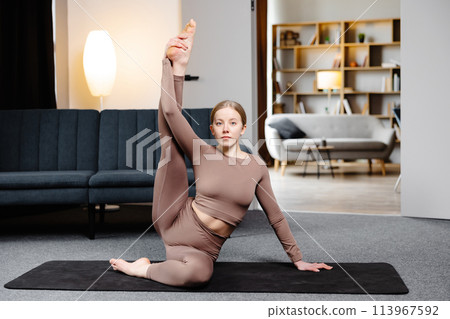 Young sporty woman practicing yoga, doing Compass pose, wearing sportswear pants and top sitting on the floor in living room 113967592