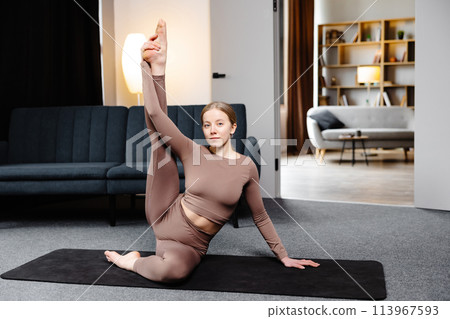 Young sporty woman practicing yoga, doing Compass pose, wearing sportswear pants and top sitting on the floor in living room 113967593