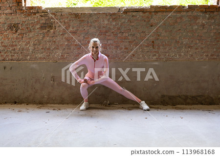 Smiling Woman in Pink Bodysuit Doing Side Lunges in Rustic Urban Studio 113968168