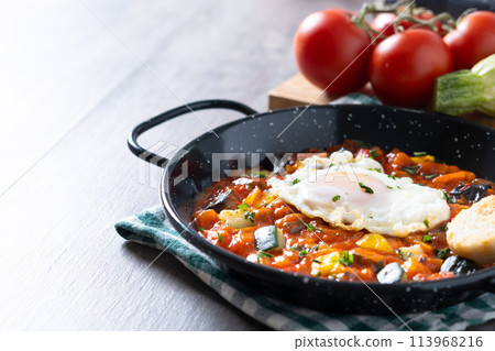 Vegetable pisto manchego with tomatoes, zucchini, peppers, onions,eggplant and egg, served in frying pan on wooden table Vegetable pisto manchego with tomatoes, zucchini, peppers, onions,eggplant and egg, served in frying pan on wooden table 113968216