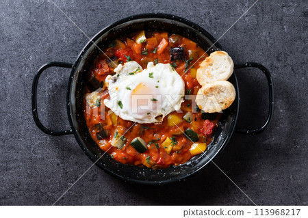 Vegetable pisto manchego with tomatoes, zucchini, peppers, onions,eggplant and egg, served in frying pan on black slate background Vegetable pisto manchego with tomatoes, zucchini, peppers, onions,eggplant and egg, served in frying pan on black slate background 113968217