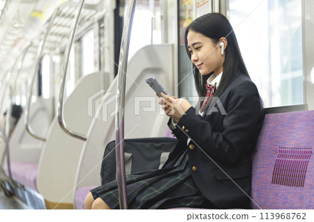 Image of a high school girl wearing earphones looking at the screen of a smartphone on the train commuting to school by train Image of a high school girl wearing earphones looking at the screen of a smartphone on the train commuting to school by train 113968762