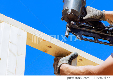 Worker in holding plank installing it on household construction using air hammer in nailing wooden beams. 113969136
