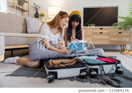 Young Asian woman packing clothes to the suitcase. Preparation for the summertime vacation. Two women are planning a trip and helping to prepare luggage to travel Young Asian woman packing clothes to the suitcase. Preparation for the summertime vacation. Two women are planning a trip and helping to prepare luggage to travel 113969176