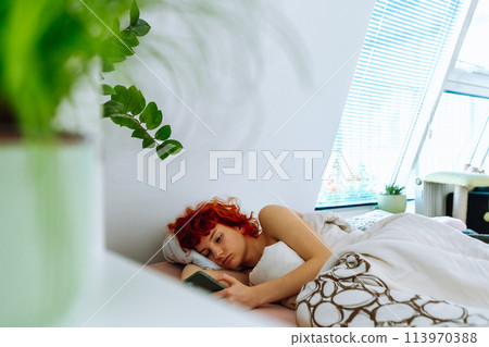 young red-haired woman lies in bed under blanket, uses smartphone, morning vacation concept young red-haired woman lies in bed under blanket, uses smartphone, morning vacation concept 113970388