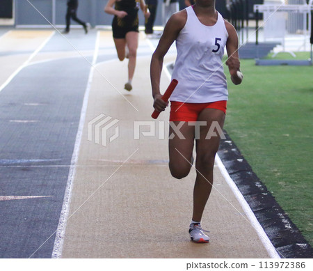 Runner racing in a relay race on an indoor track holding a baton 113972386