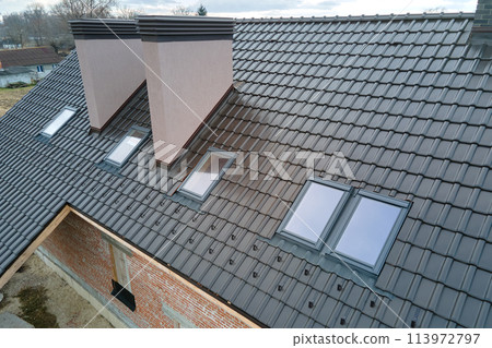 Closeup of attic windows and brick chimneys on house roof top covered with ceramic shingles. Tiled covering of building Closeup of attic windows and brick chimneys on house roof top covered with ceramic shingles. Tiled covering of building 113972797