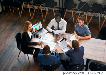 Multiracial team at office negotiation table led by female CEO. Businesswoman analyzes chart on laptop, employees discuss strategy. Corporate meeting in modern workspace, diverse group collaboration. 113972934