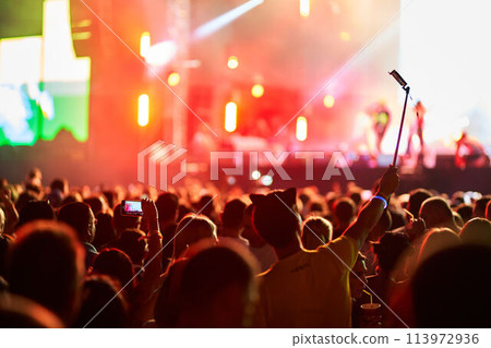 Music festival atmosphere with vibrant stage lights, enthusiastic audience enjoying live performance, fans capturing moments on smartphones, friends celebrating together in open-air venue at dusk. Music festival atmosphere with vibrant stage lights, enthusiastic audience enjoying live performance, fans capturing moments on smartphones, friends celebrating together in open-air venue at dusk. 113972936