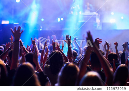 Concertgoers raise hands in rhythm at outdoor music festival, colorful stage lights illuminate excited audience. Fans enjoy live performance, nightlife energy, summer event brings people together. 113972939