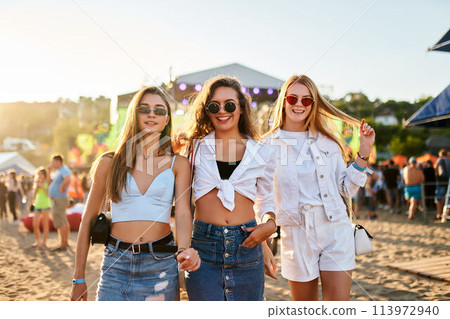 Three smiling young women walk hand in hand, sunny beach music festival. Friends in summer outfits with sunglasses, dance, socialize in coastal event. Youth culture, music on sandy shore. 113972940